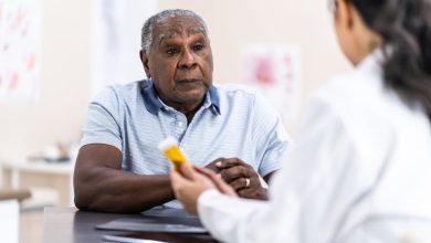 An elderly African American man sits at a desk in front of his female doctor. The doctor is holding a pill bottle and explaingin the details of the man's prescription. She is wearing a white lab coat and has long black hair pulled back into a ponytail. The elderly man is wearing casual clothing and is looking at the doctor with intent.