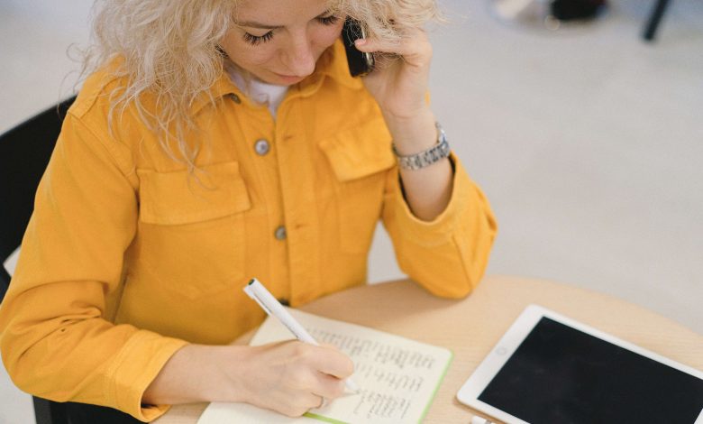 A woman in a yellow shirt is writing something on a notebook.